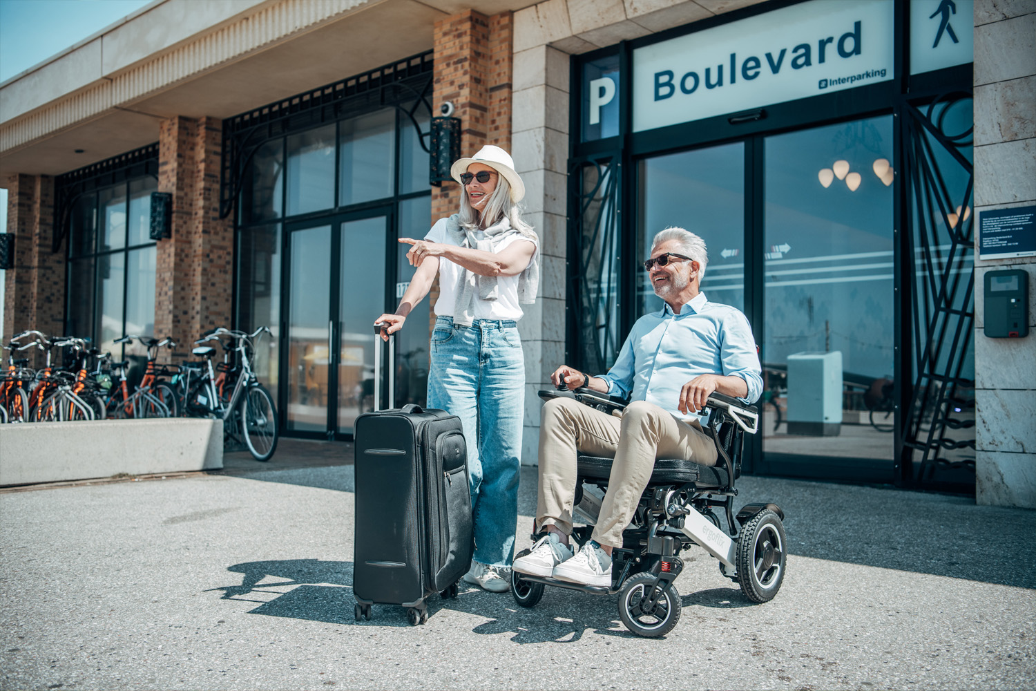 Das Bild zeigt einen Mann im Rollstuhl, neben dem eine Frau mit Koffer steht.