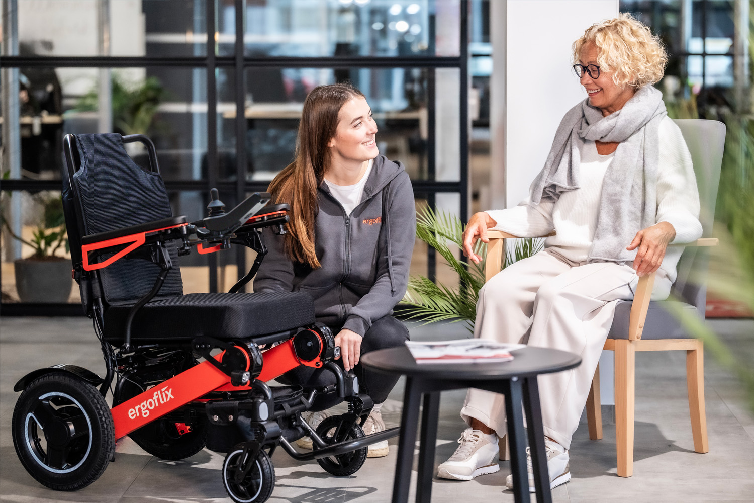 Das Bild zeigt eine Dame in einem Stuhl und eine ergoflix Mitarbeiterin, welche neben ihr kniet und sie berät. Daneben steht der ergoflix LX