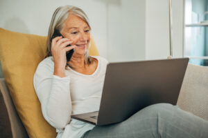 Das Bild zeigt eine &auml;ltere Frau, die auf einem Sofa sitzt, einen Laptop auf dem Schoss stehen hat und mit ihrem Handy telefoniert.