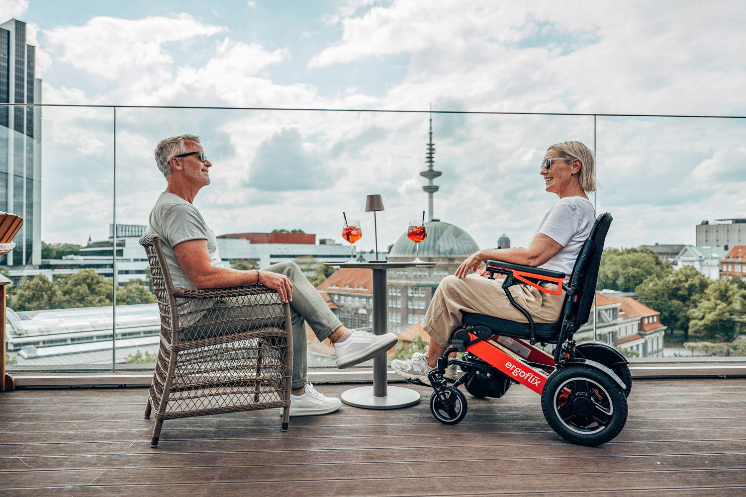 Das Bild zeigt einen Mann und eine Frau, die draußen in einem Lokal sitzen und ein Getränk trinken, sie sitzt im Rollstuhl am Tisch.