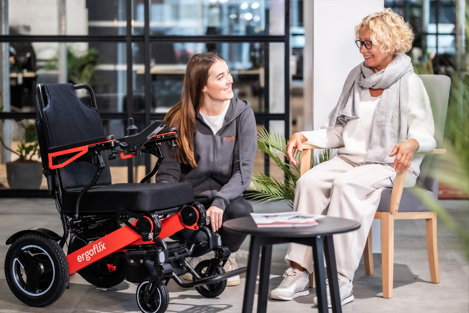 Das Foto zeigt zwei Frauen, eine davon erklärt der anderen die Funktionen eines E-Rollstuhls, der neben ihr steht.