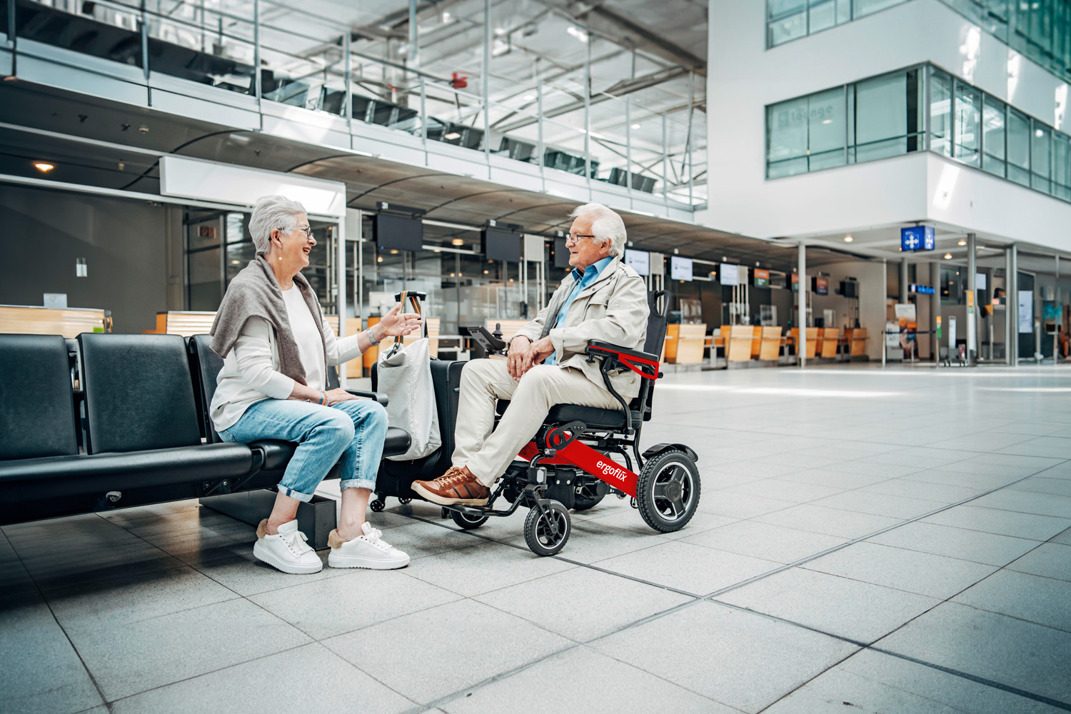 Das Bild zeigt einen Mann im Rollstuhl mit einer Frau neben sich - beide befinden sich im Innenbereich eines Flughafens.