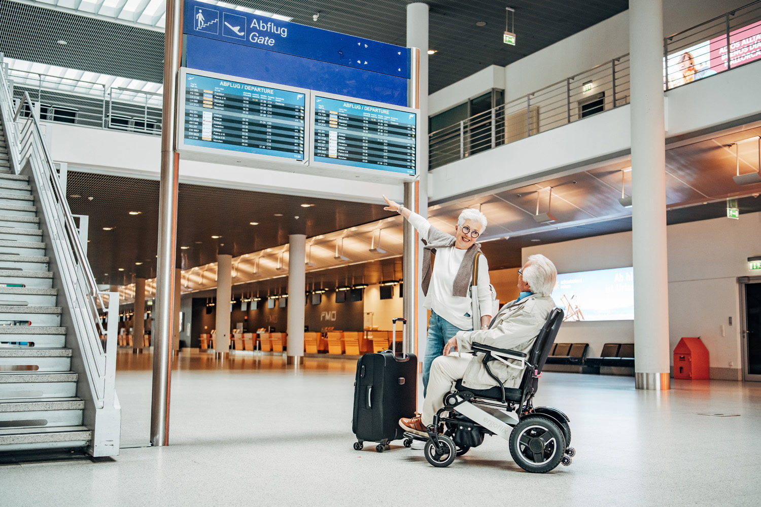 Das Bild zeigt einen Mann im Rollstuhl mit einer Frau neben sich - beide befinden sich im Innenbereich eines Flughafens.