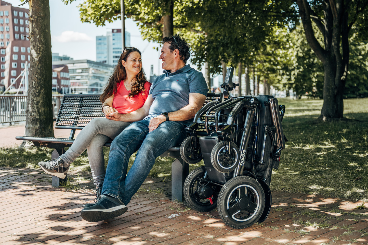 Das Bild zeigt ein Paar, welches auf einer Parkbank unter Bäumen sitzt, neben ihnen steht ein zusammengefalteter ergoflix-Rollstuhl.