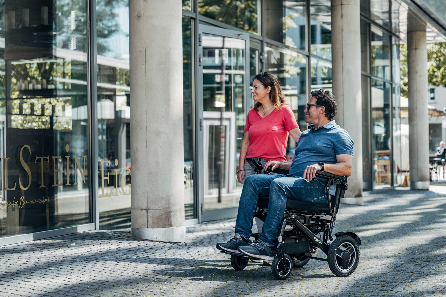 Das Bild zeigt ein Paar auf einem Ausflug, er sitzt im Rollstuhl, sie läuft daneben.