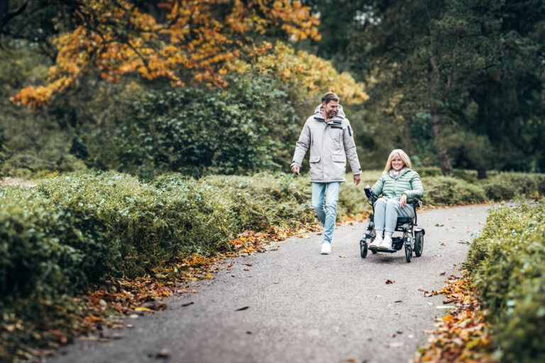 Das Bild zeigt eine Frau, die drau&szlig;en auf einem Weg mit einem Rollstuhl f&auml;hrt, neben ihr l&auml;uft ein Mann.