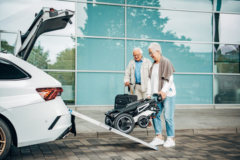 Das Bild zeigt ein &auml;lteres Paar, das einen elektrischen Faltrollstuhl &uuml;ber eine Teleskoprampe in den Kofferraum eines Autos transportiert.