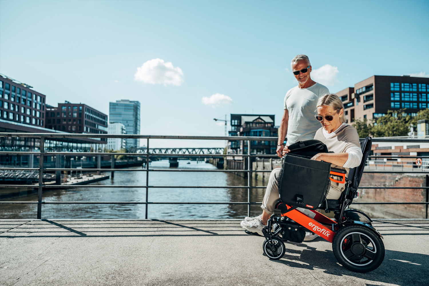 Das Bild zeigt eine Frau, die in einem ergoflix sitzt und zu ihrer linken Seite in einer Rollstuhltasche etwas sucht, neben ihr steht ein Mann.
