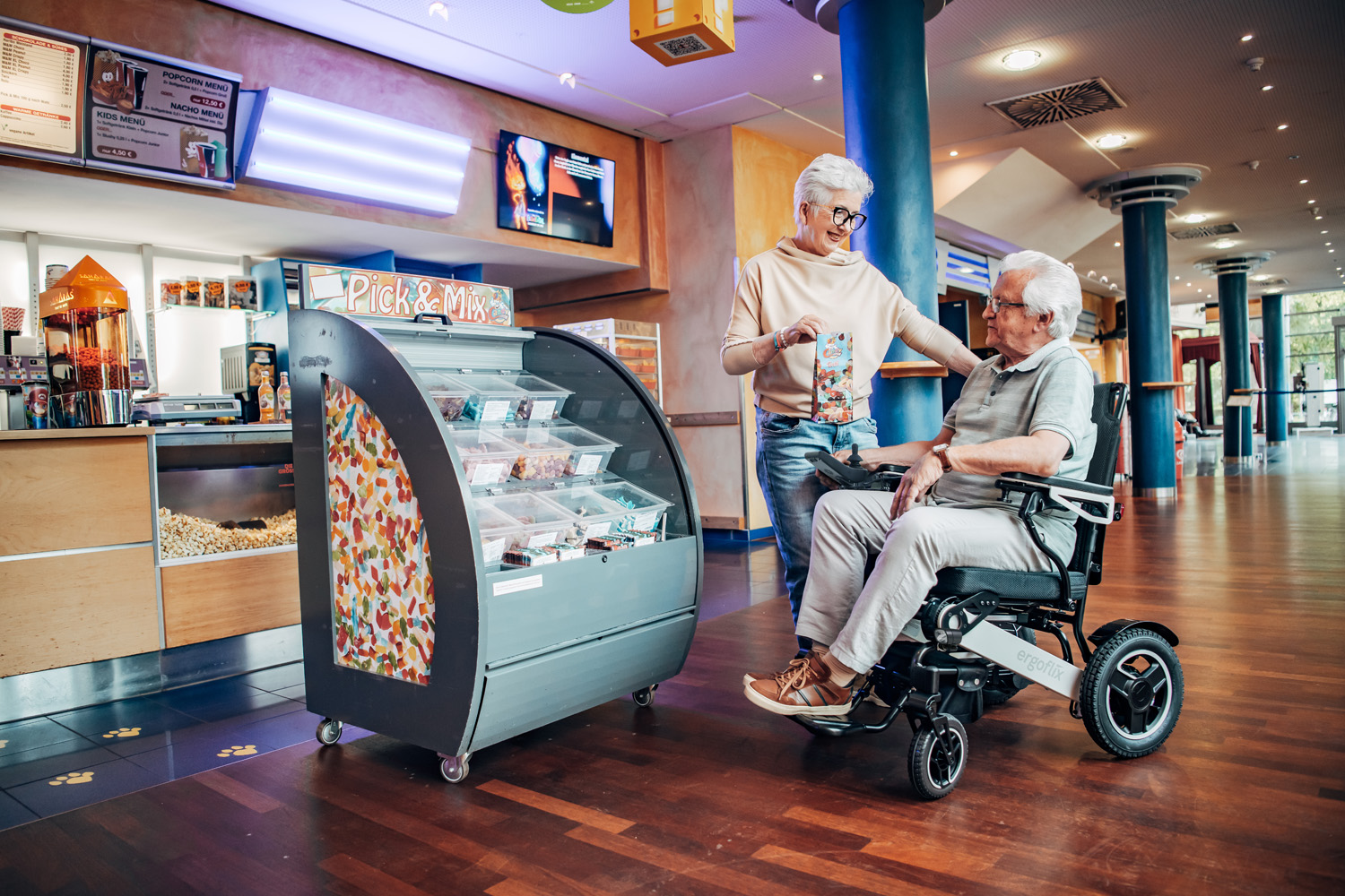 Das Bild zeigt einen Mann in einem ergoflix-Rollstuhl, neben ihm steht eine Frau und beide befinden sich an einer Kino-Theke.