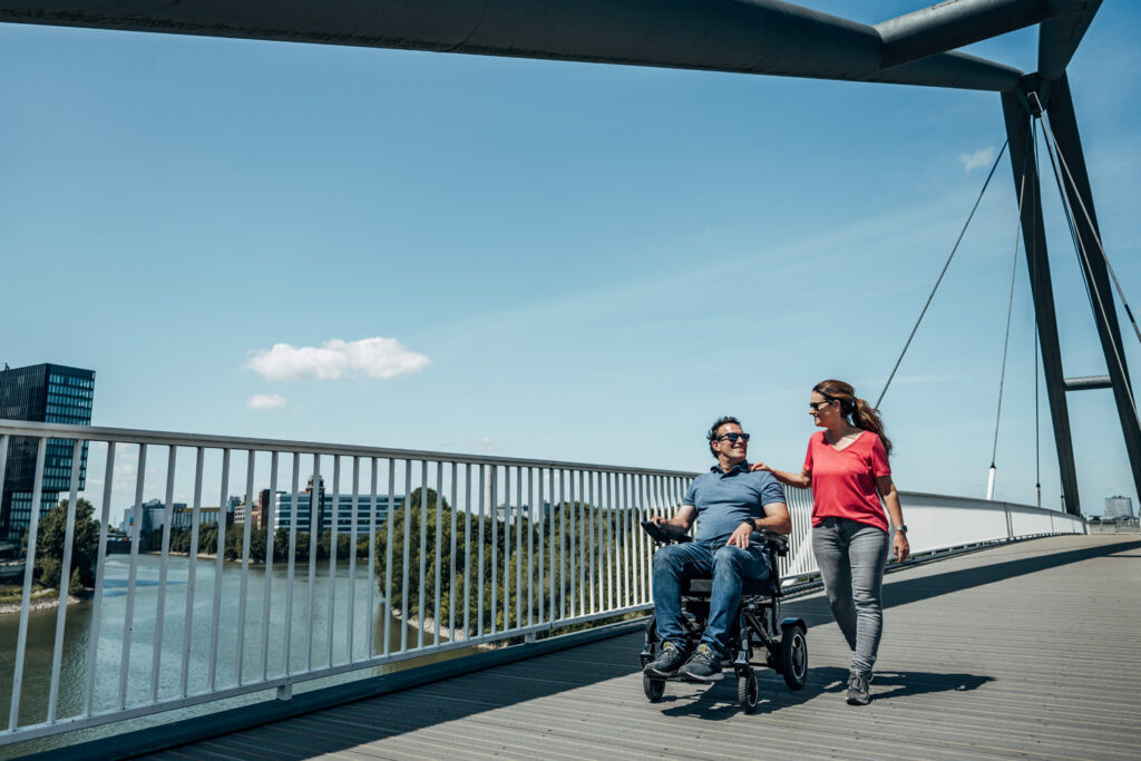 Das Bild zeigt ein Paar auf einem Ausflug, er sitzt im Rollstuhl, sie läuft daneben.