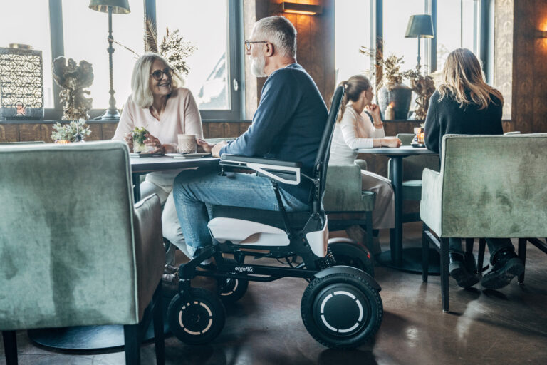 Das Bild zeigt eine Szenerie in einem Restaurant, im Vordergrund sitzt eine Frau am Tisch, ein Mann sitzt neben ihr am Tisch in einem Elektrorollstuhl.