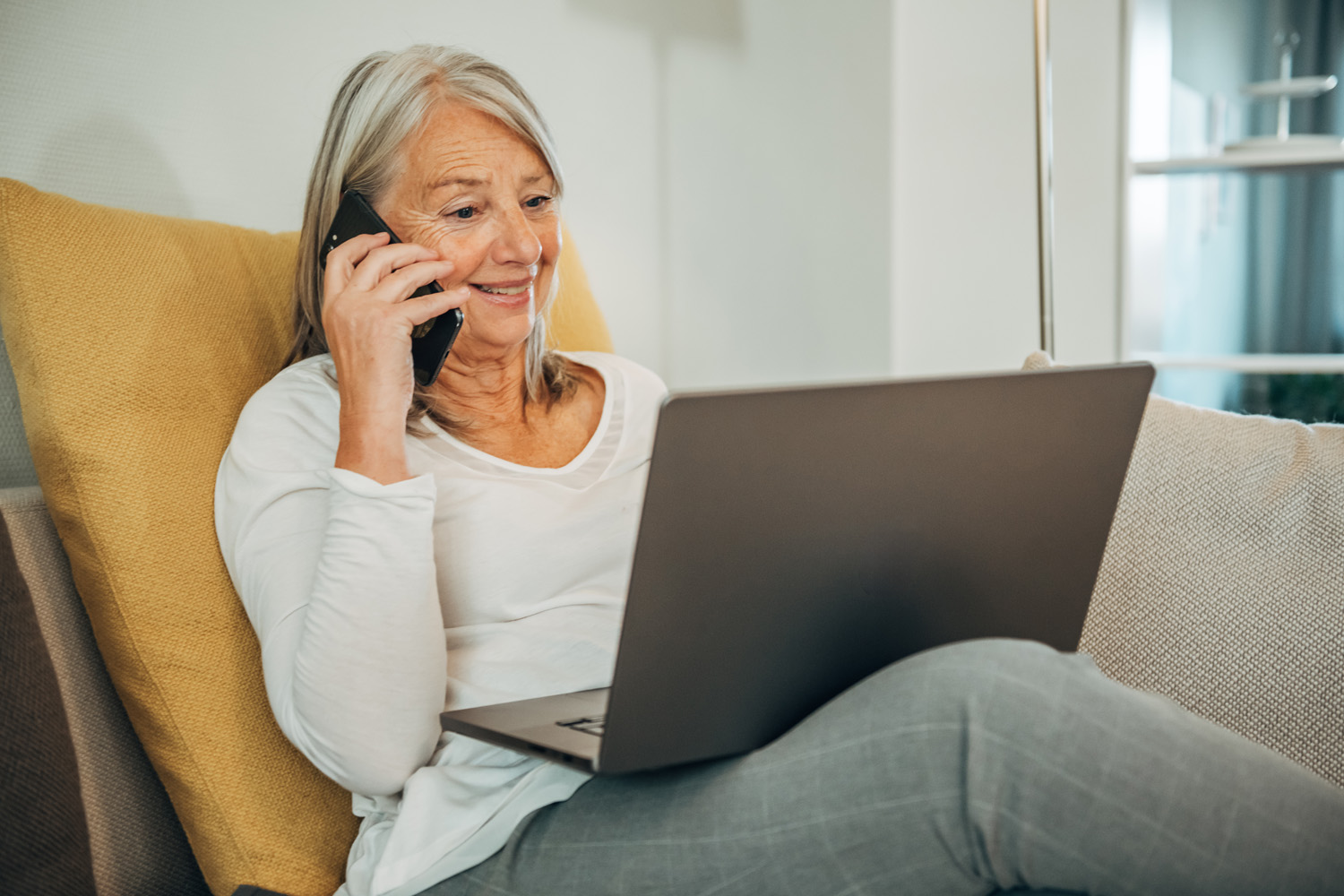 Das Bild zeigt eine Frau, die auf dem Sofa sitzt, telefoniert und einen Laptop auf dem Schoß liegen hat.