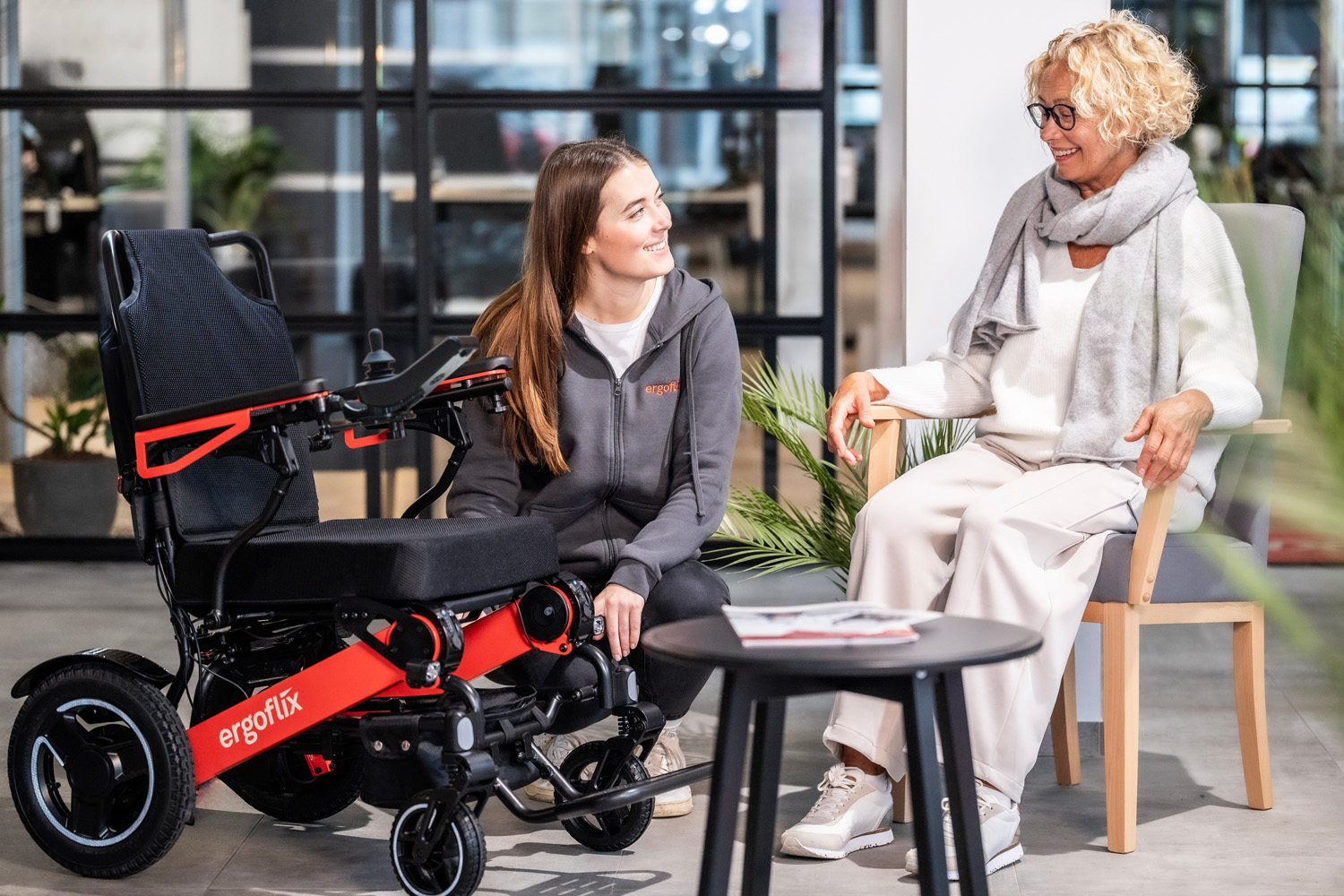 Das Bild zwei Frauen, eine sitzt auf einem Stuhl, die andere kniet neben einem ergoflix® LX und erklärt der anderen Frau etwas.