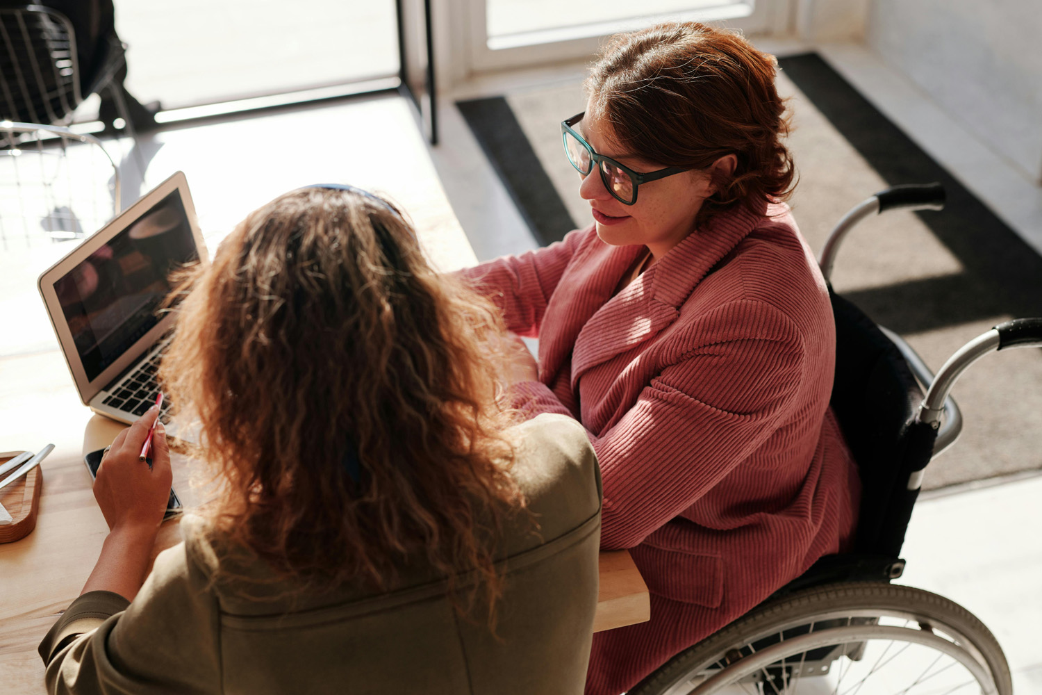 Das Bild zeigt zwei Frauen, die nebeneinander am Tisch vor einem Laptop sitzen, eine der Frauen sitzt in einem manuellen Rollstuhl.