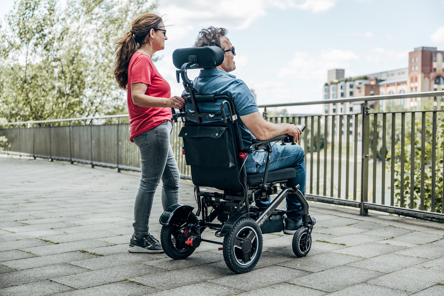 Das Bild zeigt ein Paar von hinten, der Mann sitzt in einem Rollstuhl mit Kopfstütze, die Frau steht daneben.