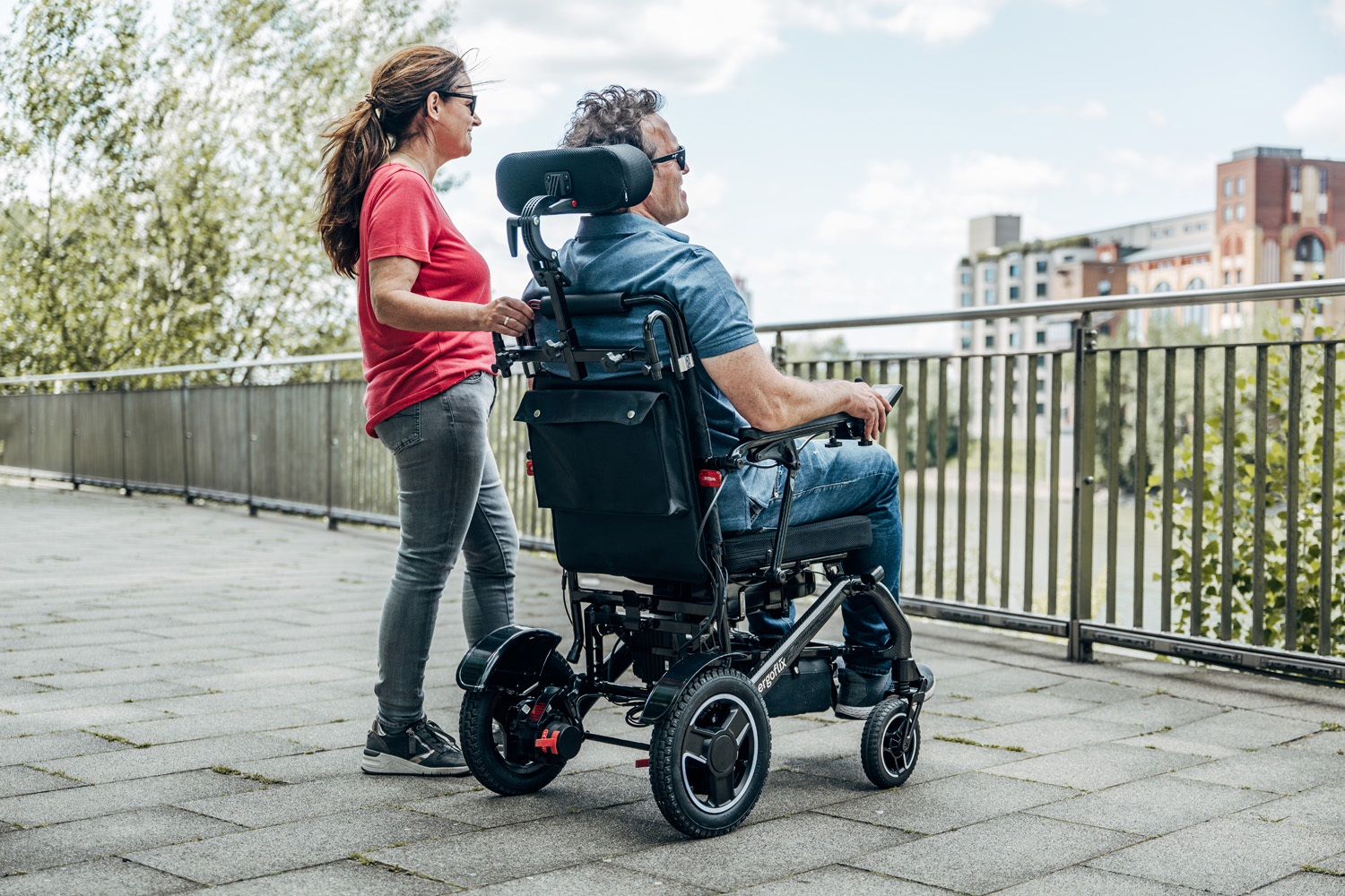 Das Bild zeigt ein Paar von hinten, der Mann sitzt in einem Rollstuhl mit Kopfstütze, die Frau steht daneben.