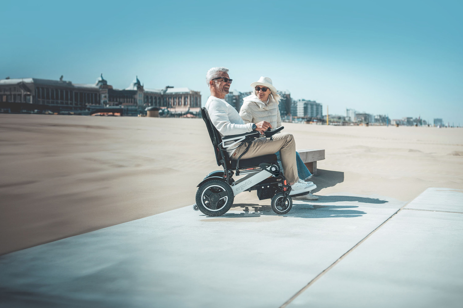 Das Bild zeigt einen Mann im ergoflix-Rollstuhl an einer Strandpromenade, neben ihm sitzt eine Frau.