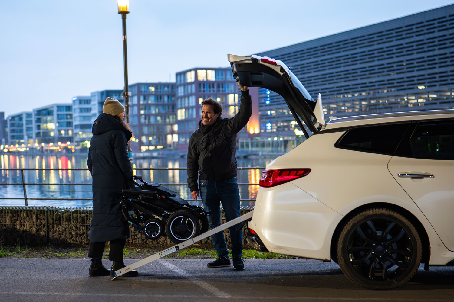 Das Bild zeigt eine Frau, die über eine Rampe einen Rollstuhl in ein Auto verlädt, ein Mann steht daneben.