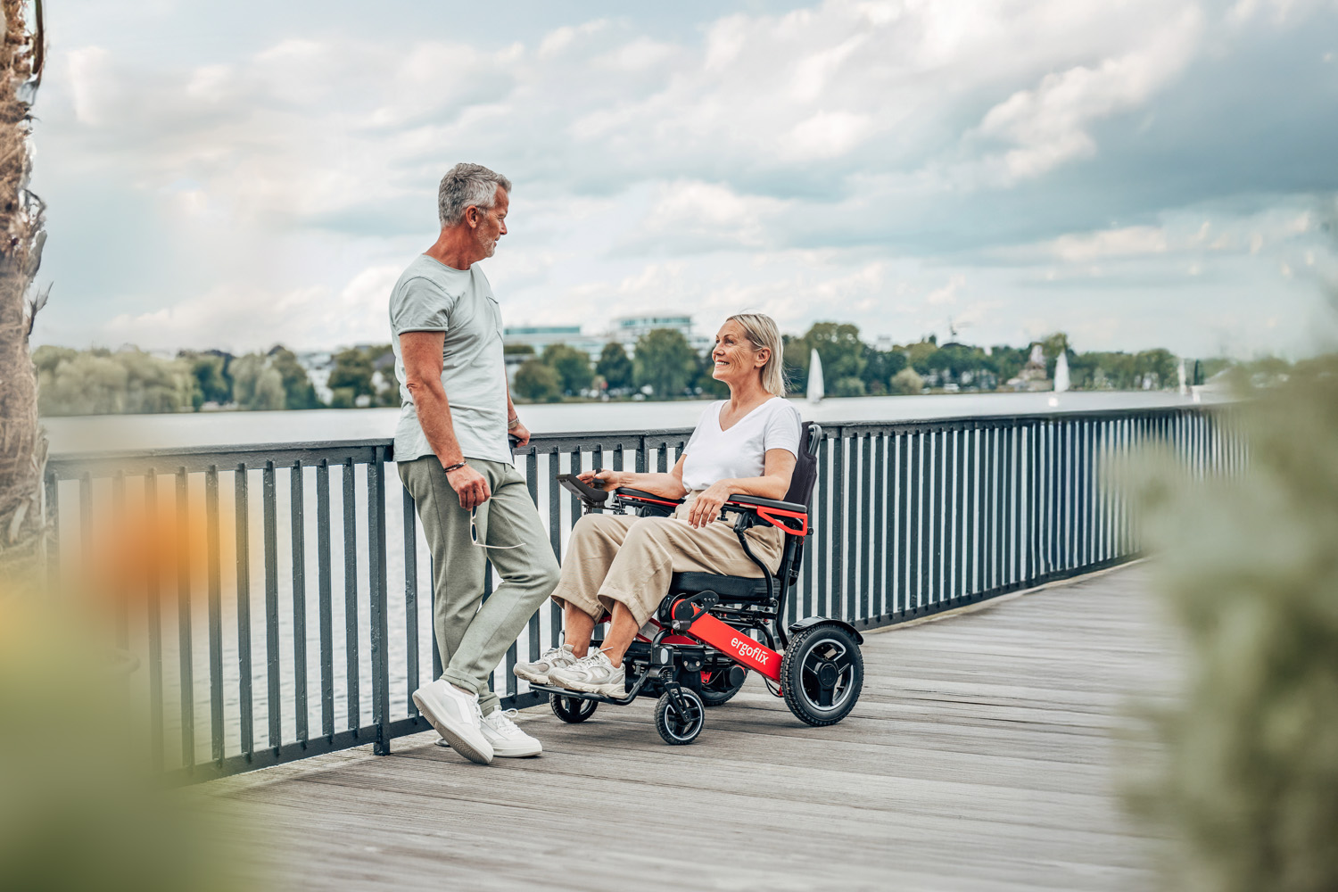 Das Bild zeigt ein Pärchen, sie sitzt im Rollstuhl, er steht ihr gegenüber und lehnt sich an ein Geländer, dahinter befindet sich ein Gewässer.