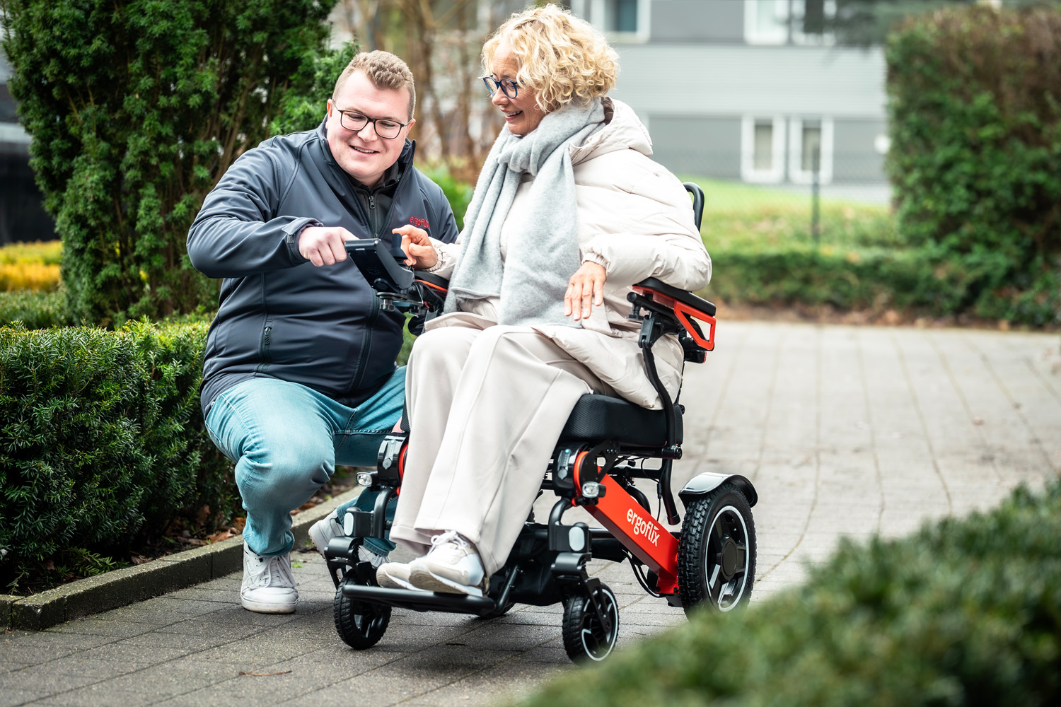 Das Foto zeigt eine Frau sitzend im Rollstuhl, während ein Mann daneben ihr etwas zum Rollstuhl erklärt.