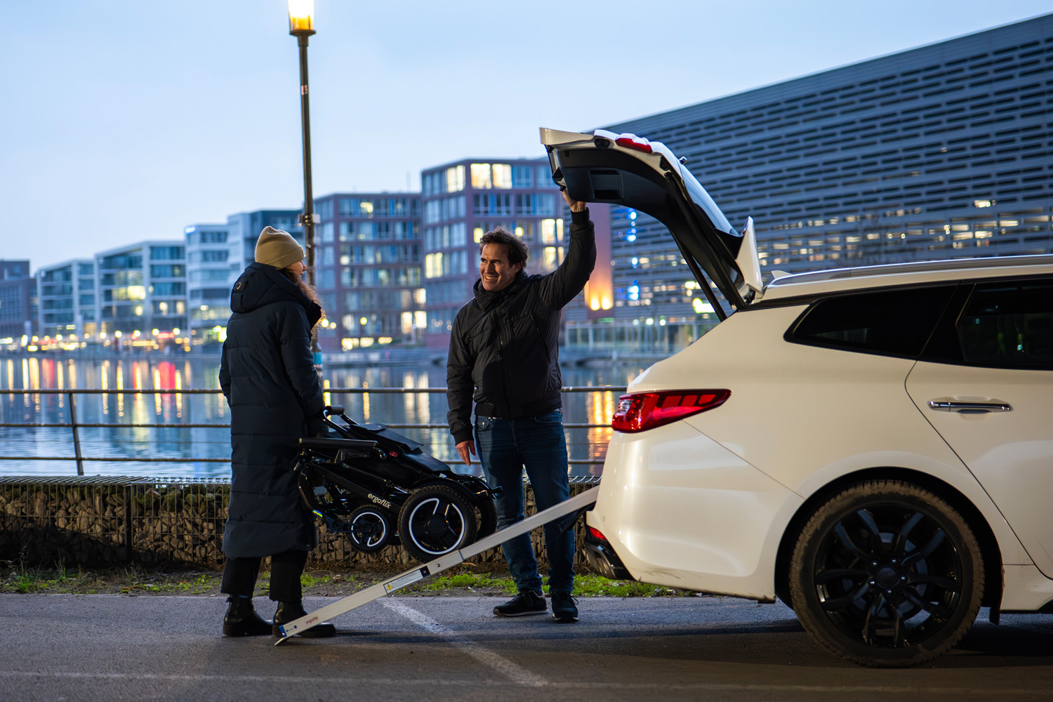 Das Bild zeigt einen Mann und eine Frau, die über eine Rampe einen Rollstuhl in den Autokofferraum verladen.