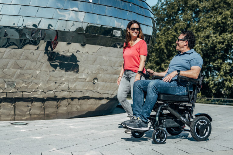Das Bild zeigt einen Rollstuhlfahrer, der &uuml;ber einen &ouml;ffentlichen Platz f&auml;hrt, neben ihm l&auml;uft eine Frau.