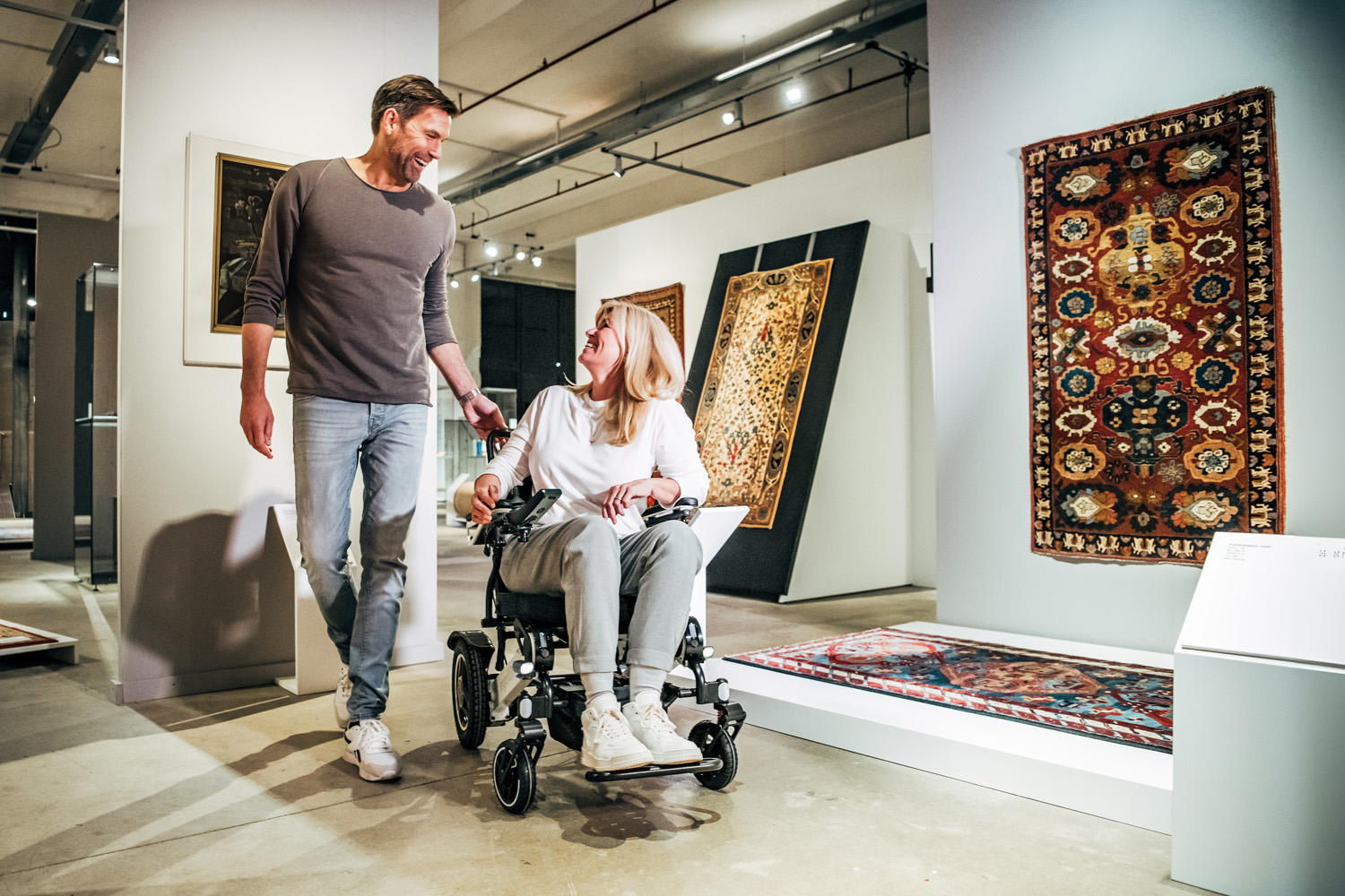 Das Bild zeigt eine Frau, die im Rollstuhl sitzend durch ein Textilmuseum fährt, neben ihr läuft ein Mann.