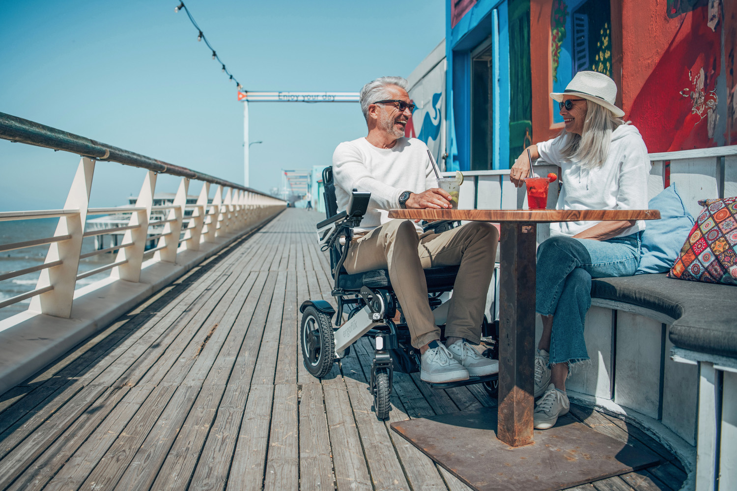 Das Bild zeigt einen Mann im Rollstuhl, neben ihm sitzt eine Frau und beide befinden sich im Außenbereich eines Lokals.