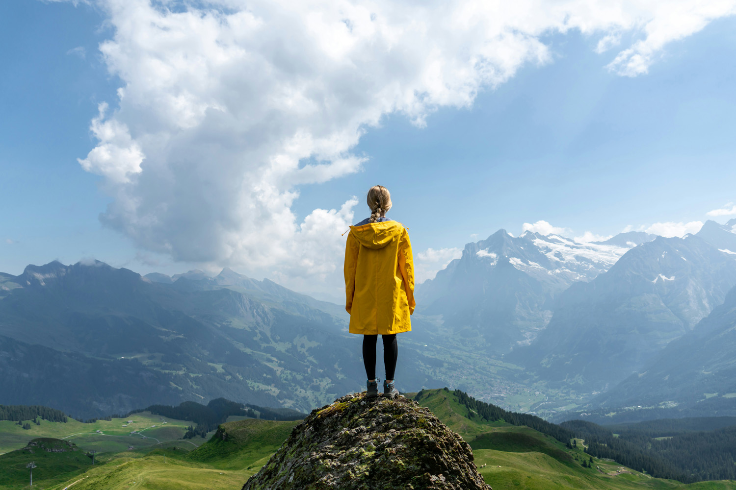 Auf dem Bild ist eine Berglandschaft zu sehen, im Vordergrund steht mittig eine Frau auf einem kleinen Felsen, sie ist von hinten zu sehen.