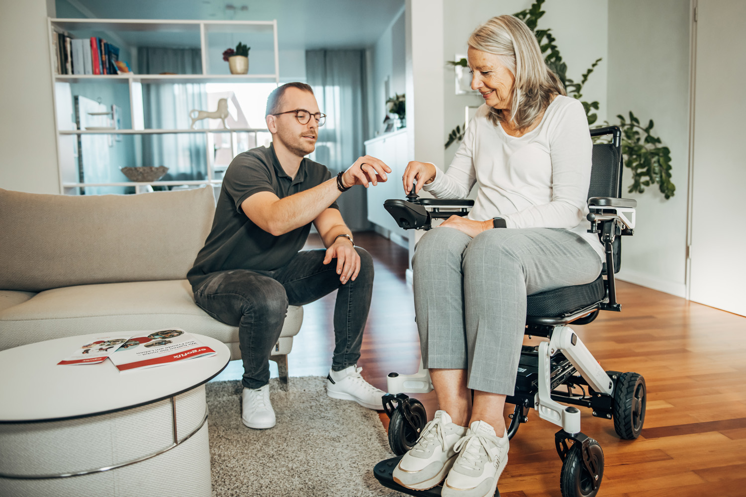 Das Foto zeigt eine Frau sitzend im Rollstuhl in einer Wohnung, während ein Mann daneben ihr etwas zum Rollstuhl erklärt.