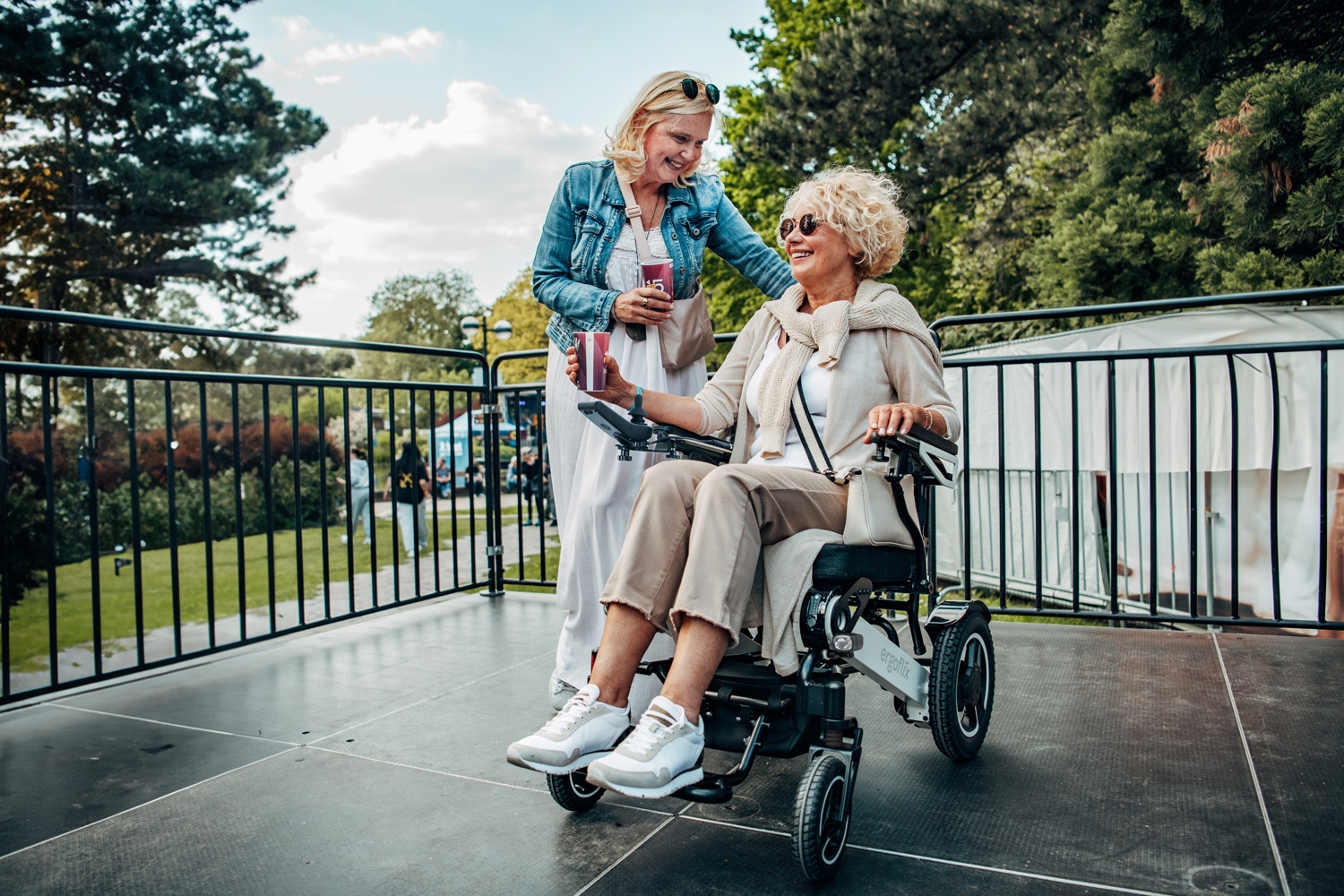 Das Bild zeigt eine Frau im Rollstuhl, neben ihr steht eine Frau und beide befinden sich auf einem Festival.