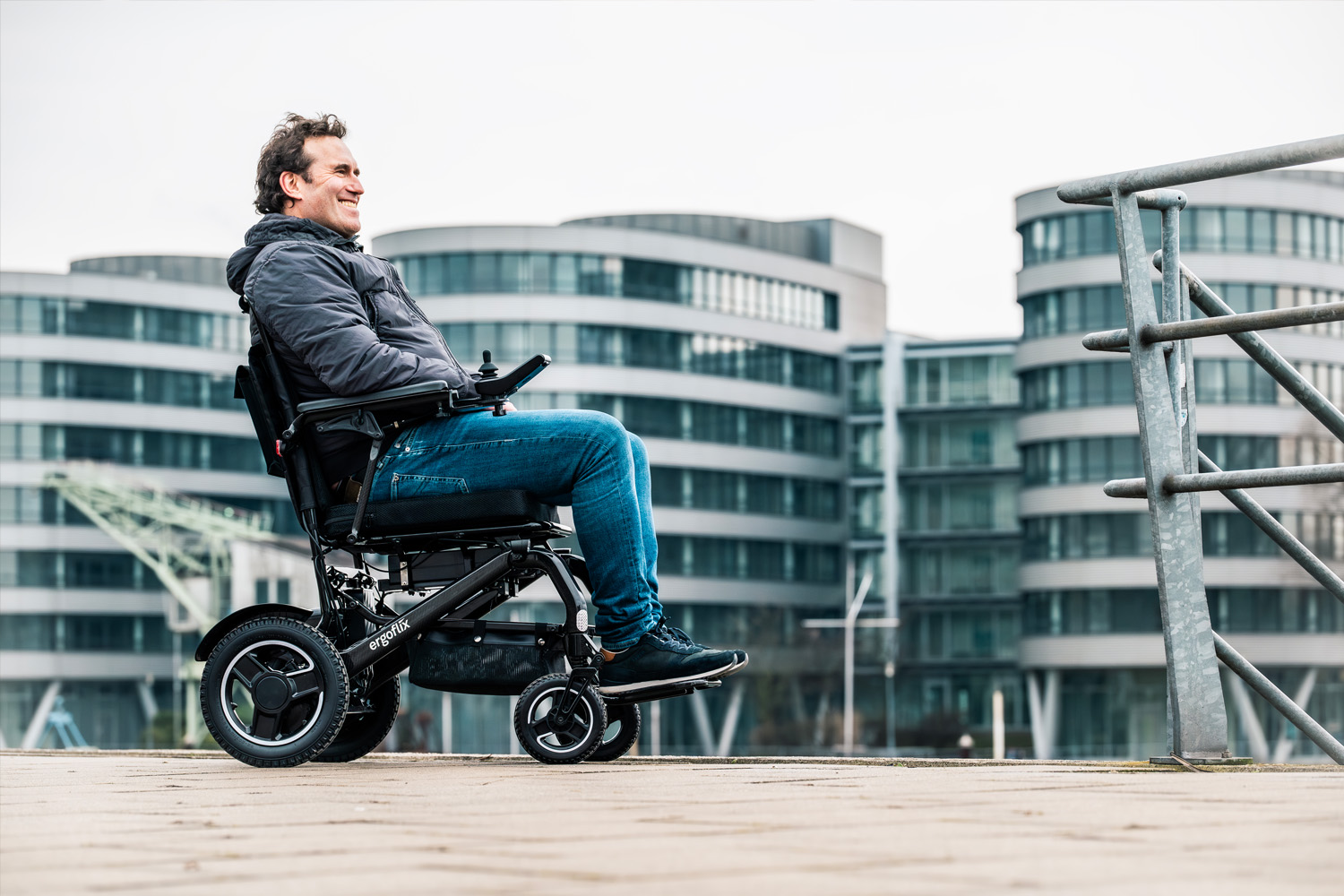 Das Bild zeigt einen Mann, der mit großen Gebäuden im Hintergrund in einem Elektrorollstuhl sitzt.