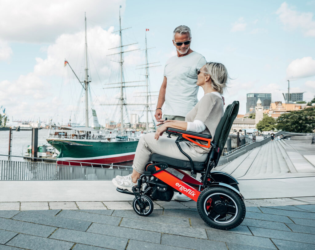 Das Bild zeigt einen Mann und eine Frau am Hafen, sie sitzt in einem ergoflix-Rollstuhl und er steht neben ihr.