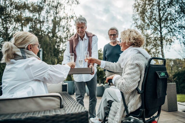 Dieses Bild zeigt eine Gruppe an Personen, die mit Sekt ansto&szlig;en. Eine Frau sitzt im Rollstuhl,
