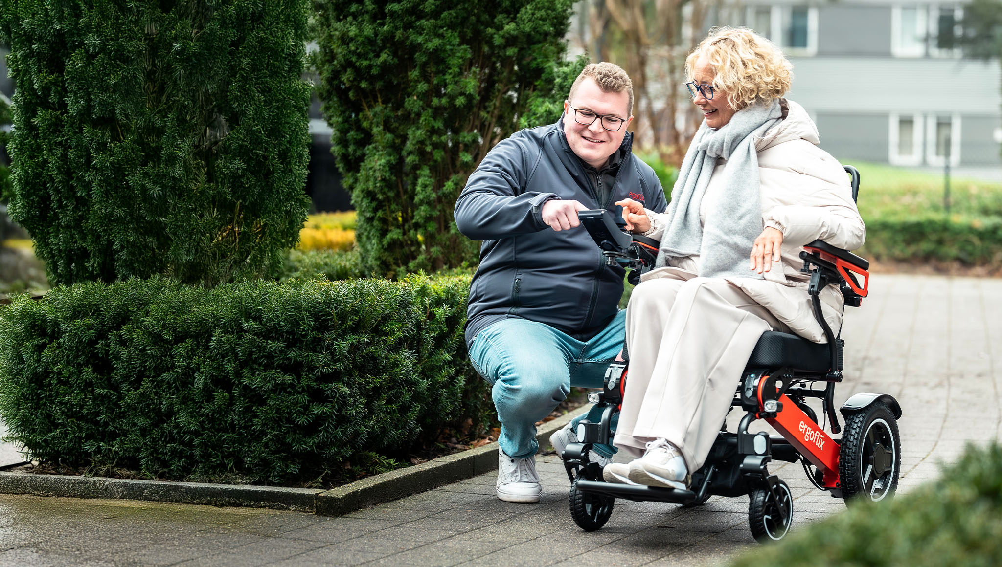 Junger Mann erklärt einer Dame das Bedienmodul eines roten faltbaren Elektrorollstuhls im Freien bei einer Probefahrt.