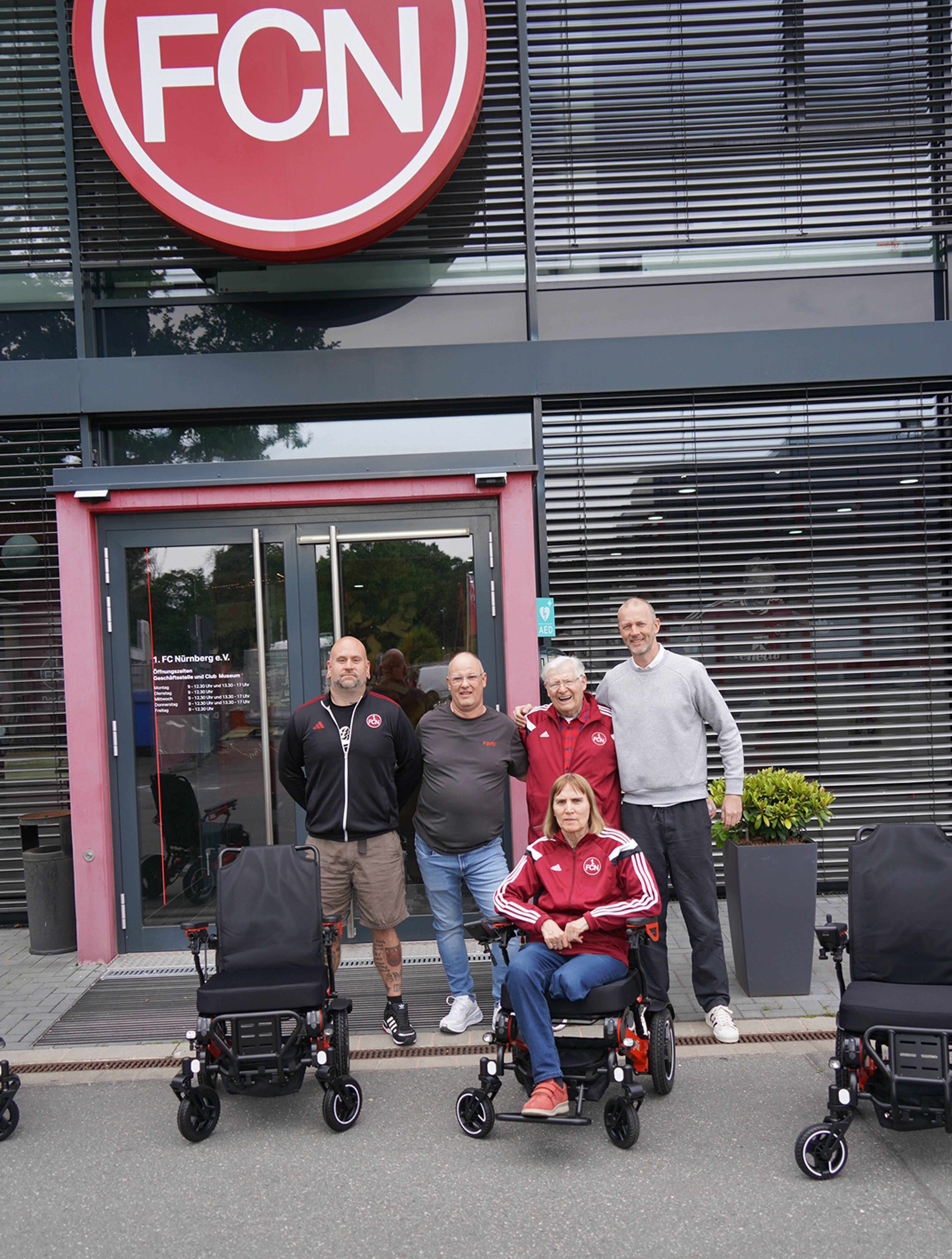 Das Bild zeigt mehrere Menschen (vom 1. FC N&uuml;rnberg und von ergoflix), die mit Rollst&uuml;hlen von ergoflix vor einem Geb&auml;ude mit dem Vereinslogo stehen.