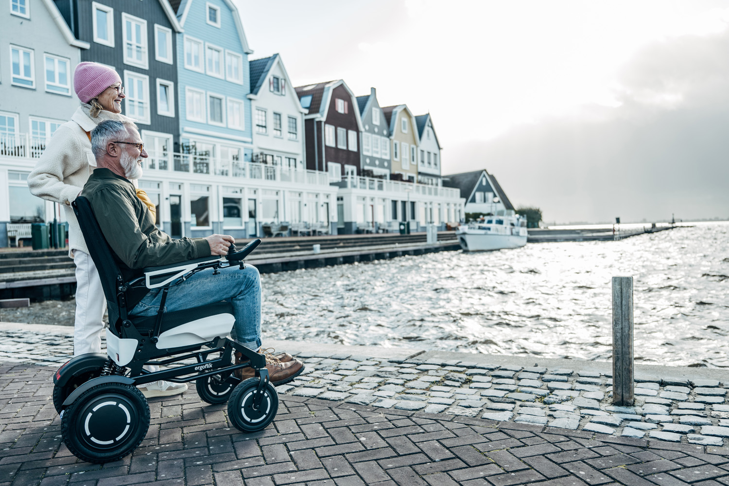 Das Bild zeigt einen Mann mit einem Handy in der Hand, er sitzt auf einem Sofa, neben ihm steht ein gefalteter Elektrorollstuhl.