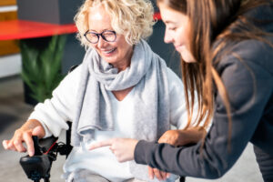 Das Foto zeigt zwei Frauen, eine davon erkl&auml;rt der anderen die Funktionen eines E-Rollstuhls, der neben ihr steht.