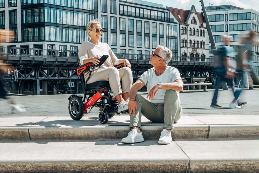 Eine ältere Dame in einem roten ergoflix® LX Rollstuhl, die sich mit einem älteren Herrn, welcher auf einer Treppe sitzt, unterhält.