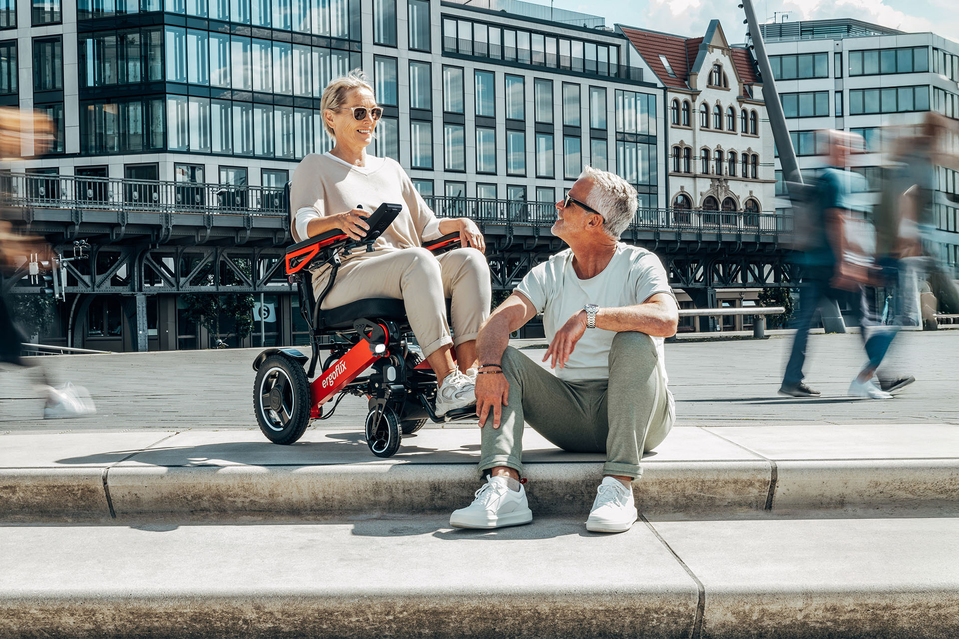 Eine ältere Dame in einem roten ergoflix® LX Rollstuhl, die sich mit einem älteren Herrn, welcher auf einer Treppe sitzt, unterhält.