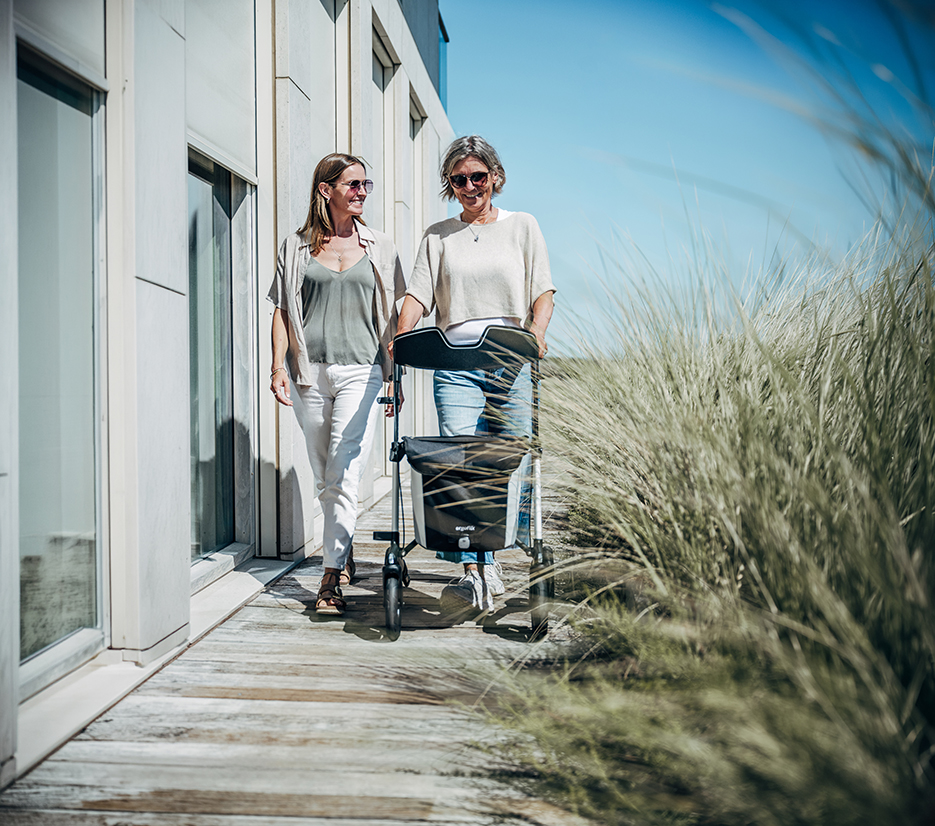 Two women walking with an ergoflix rollator along a boardwalk in the dunes